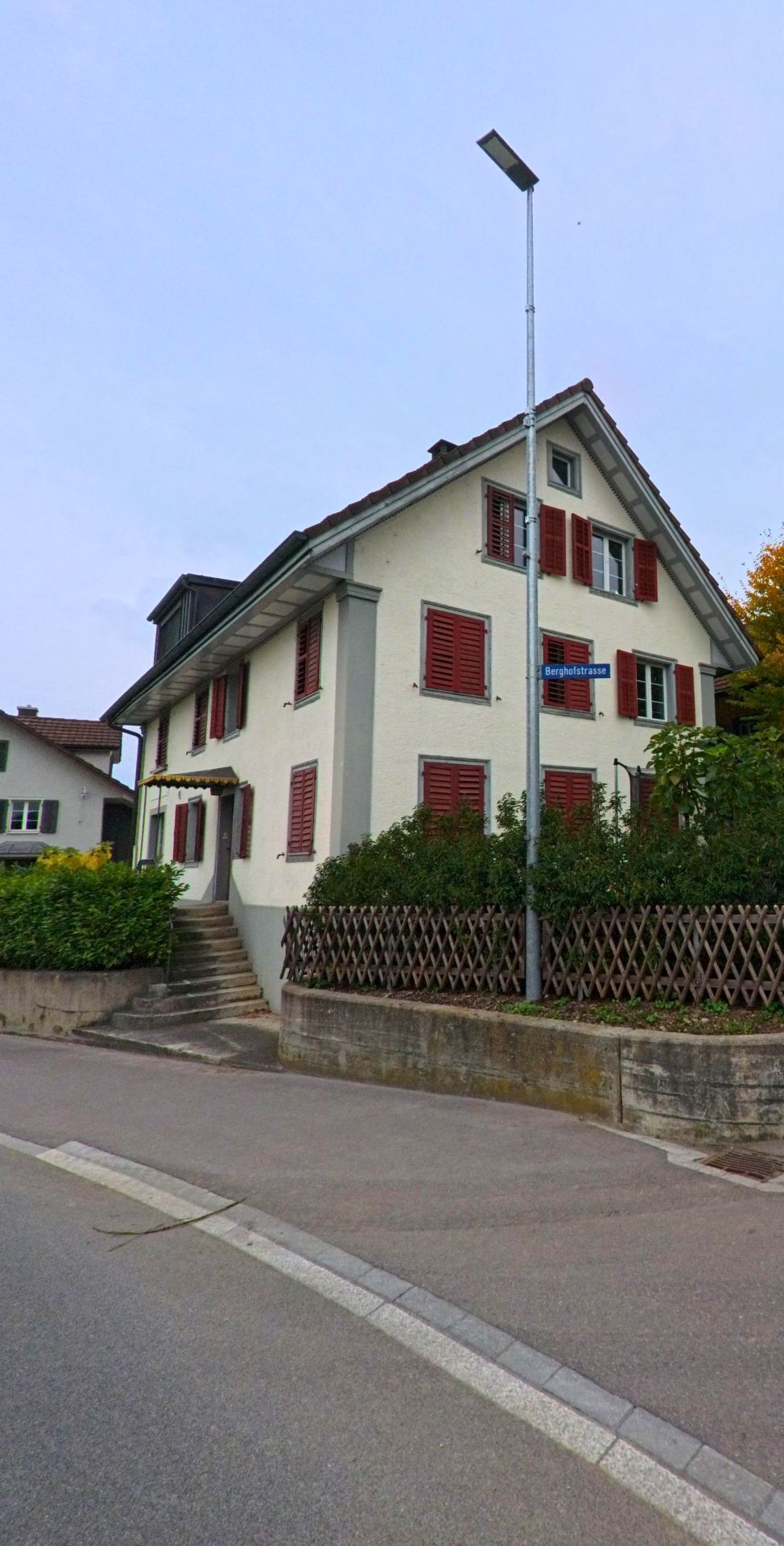In Unterlunkhofen (8918, Kanton Aargau) steht ein helles Wohnhaus mit roten Fensterlaeden an der Berghofstrasse, leicht erhoeht über dem Strassenniveau.