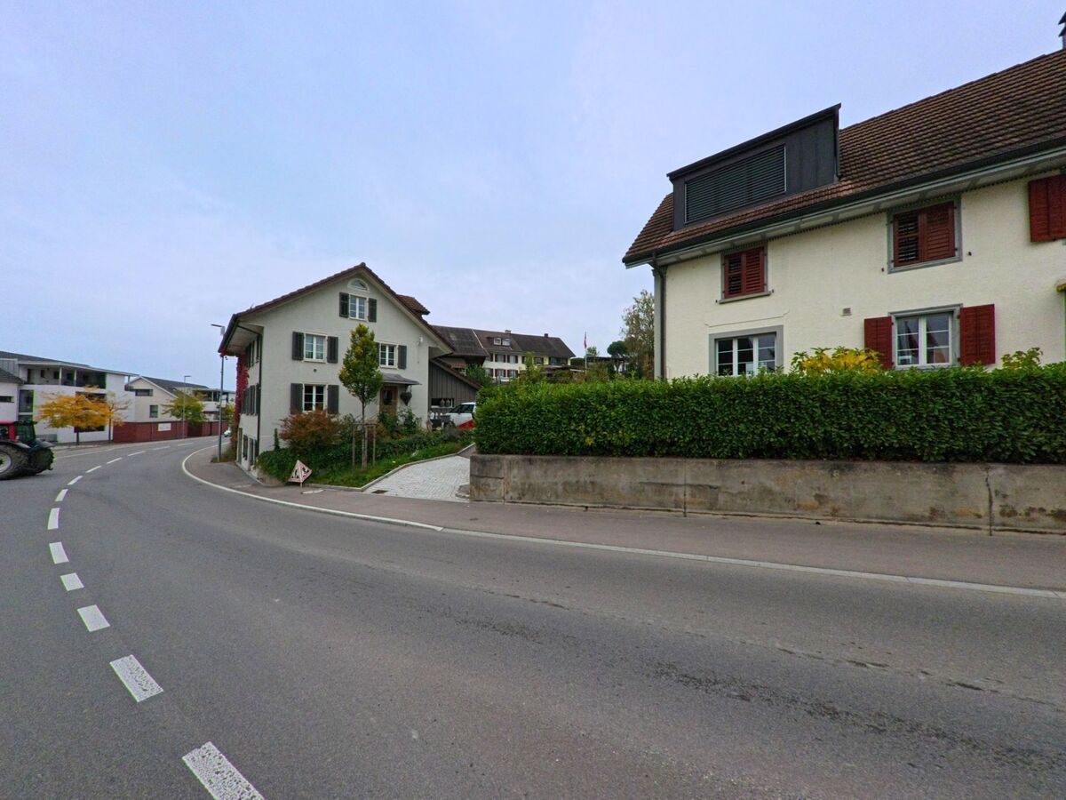 Eine sanft geschwungene Strasse in Unterlunkhofen (8918, Kanton Aargau) fuehrt an gepflegten Wohnhaeusern mit Hecken und kleinen Gaerten vorbei.