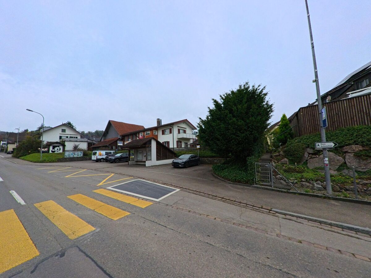 Eine Hauptstrasse in Unterlunkhofen (8918, Kanton Aargau) fuehrt an einer Bushaltestelle, mehreren Wohnhaeusern und einem bepflanzten Hang mit Treppe vorbei.