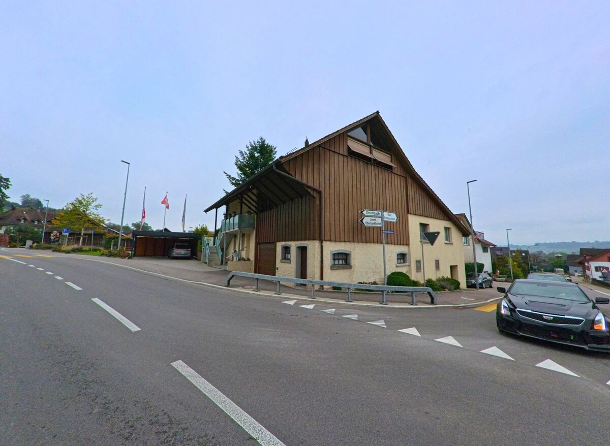 An einer Kreuzung in Unterlunkhofen (8918, Kanton Aargau) steht ein Gebaeude mit Holzfassade und Wegweisern, waehrend die Strasse leicht ansteigt.