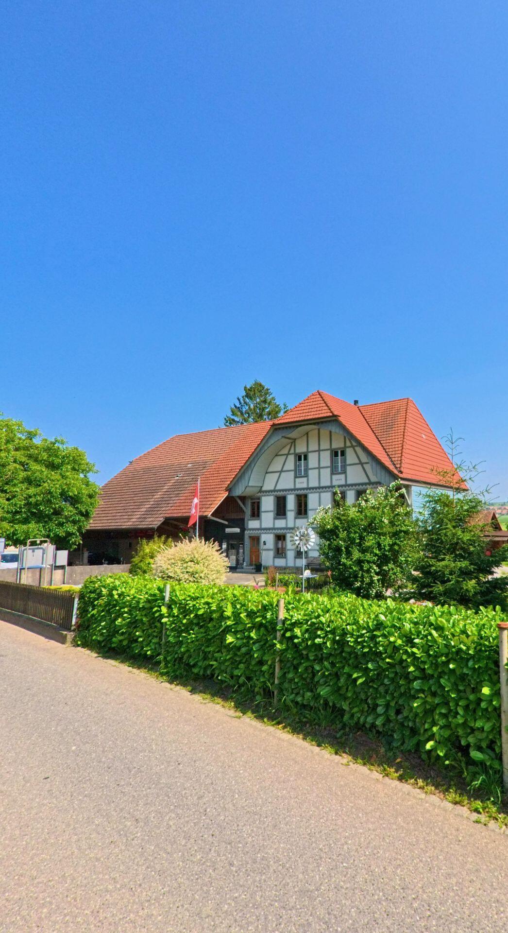Hinter einer dichten Hecke steht in Unterramsern (4588, Kanton Solothurn) ein stattliches Bauernhaus mit markanter Fassade, rotem Ziegeldach und angrenzendem Wirtschaftsbereich.