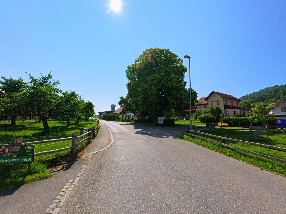 Auf einer leicht geschwungenen Dorfstrasse in Unterramsern (4588, Kanton Solothurn) führen beidseitige Holzzäune entlang von Wiesen und Bäumen durch das ländliche Siedlungsgebiet.