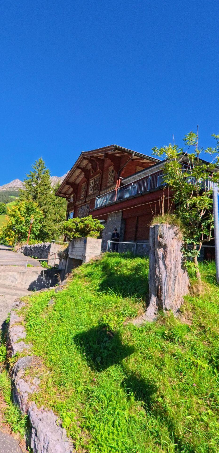 Ein traditionelles Holzhaus steht erhöht auf einer begrünten Terrasse in Unterschächen (6465, Kanton Uri), umgeben von Bäumen, Treppenwegen und alpiner Landschaft unter blauem Himmel.