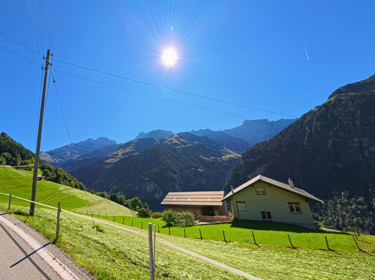 Auf einer Anhöhe liegen Wohnhäuser und gemähte Wiesen in Unterschächen (6465, Kanton Uri), während sich im Hintergrund die Bergkette unter klarem Himmel erhebt.