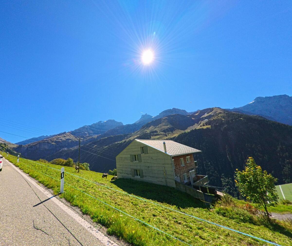 Entlang einer Hangstrasse steht ein einzelnes Wohnhaus über grünen Wiesen in Unterschächen (6465, Kanton Uri), mit freiem Blick auf die umliegenden Berge unter strahlender Sonne.