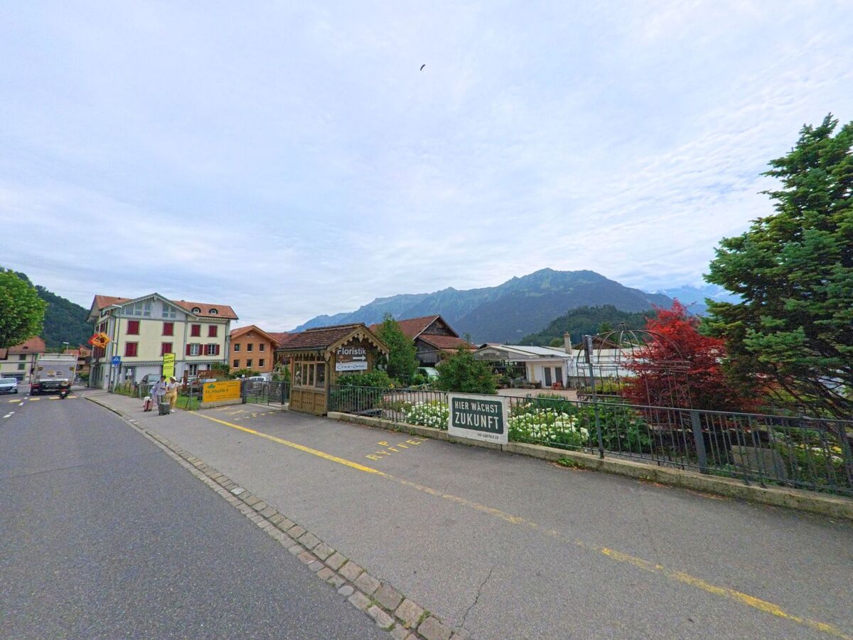 Entlang der Hauptstrasse von Unterseen (3800, Kanton Bern) liegen Gärtnerei und Wohnhäuser mit Blick auf grüne Hügel und bewaldete Berge.
