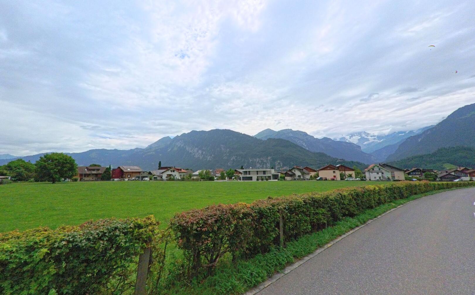 Eine Landstrasse in Unterseen (3800, Kanton Bern) führt an grünen Wiesen vorbei mit Blick auf die umliegenden Alpen unter leicht bewölktem Himmel.