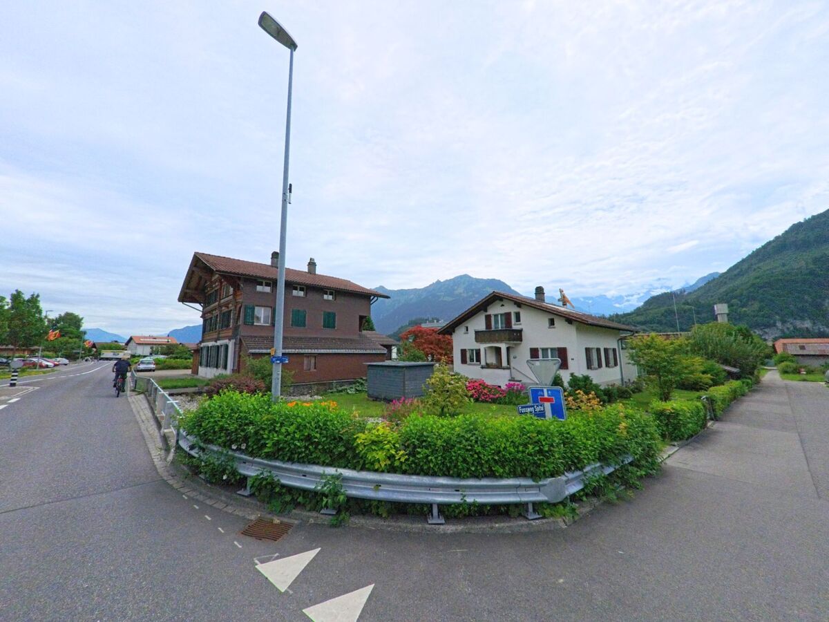 In Unterseen (3800, Kanton Bern) stehen gepflegte Wohnhäuser mit Blumengärten vor einer beeindruckenden Bergkulisse unter leicht bewölktem Himmel.