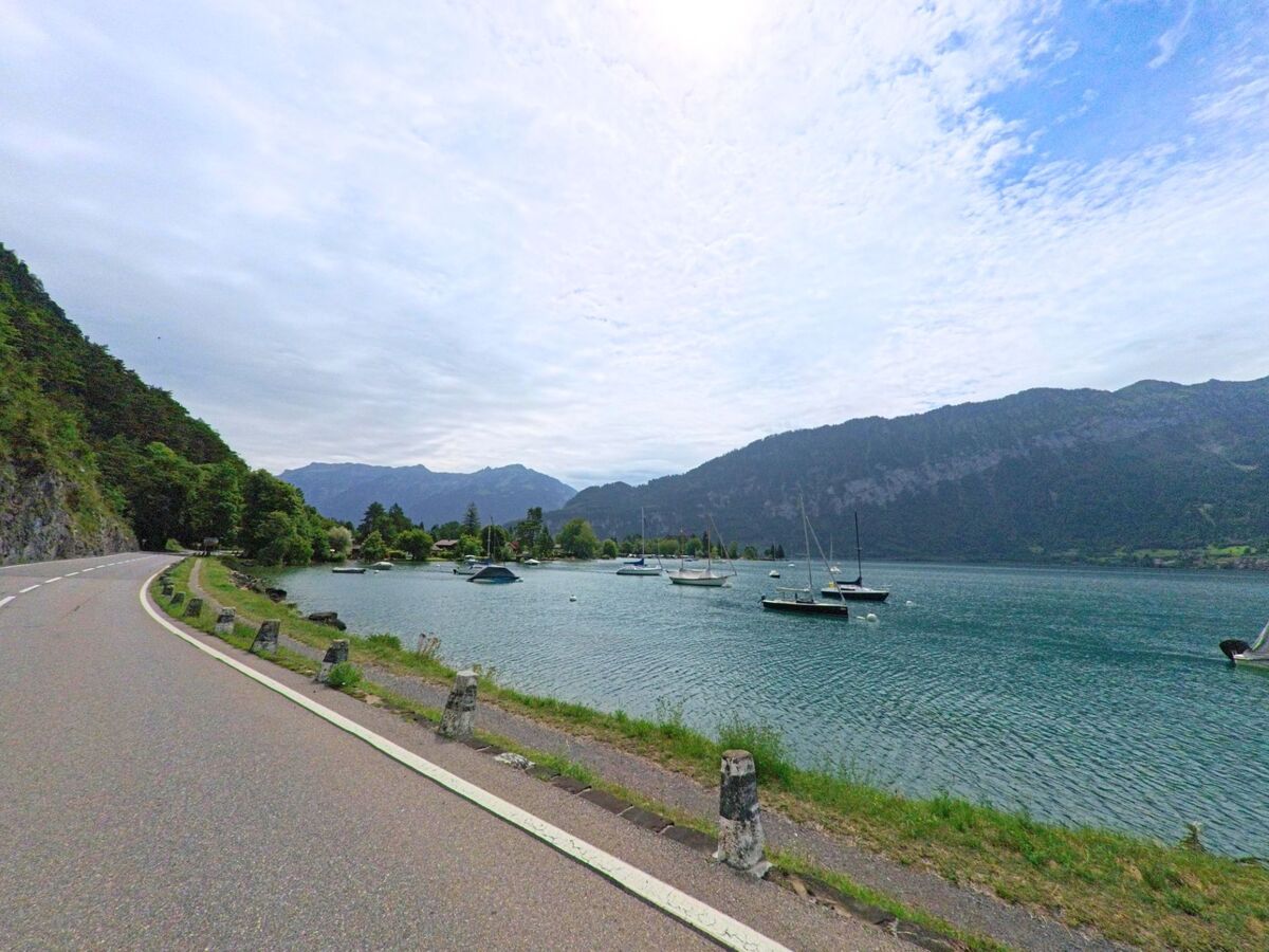 Entlang des Thunersees bei Unterseen (3803, Kanton Bern) verläuft eine kurvige Strasse neben ruhendem Wasser und Segelbooten vor Bergkulisse.