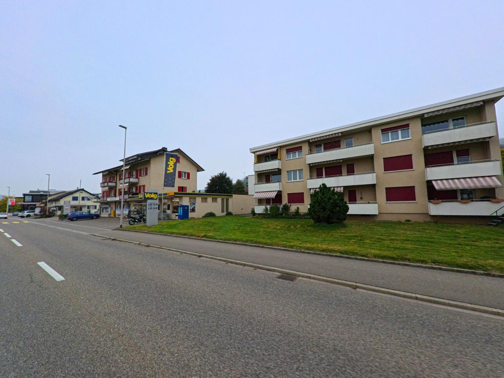 Entlang einer breiten Strasse mit Wohnbauten und einem Lebensmittelgeschäft zeigt sich das Quartierbild von Untersiggenthal (5417, Kanton Aargau) an einem ruhigen Tag.