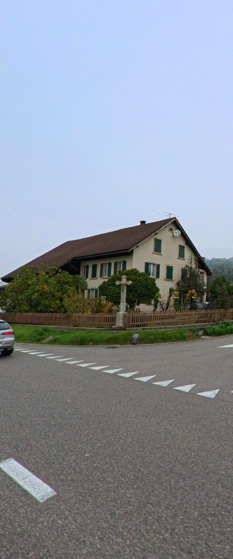 Ein älteres Wohnhaus mit grünen Fensterläden und einem steinernen Wegkreuz steht an einer Strassenecke in Untersiggenthal (5417, Kanton Aargau) in ländlicher Umgebung.