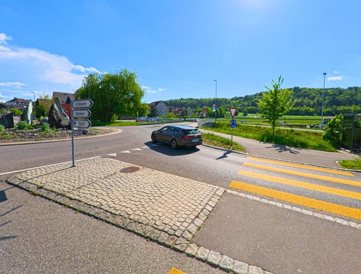 Am Ortseingang von Urdorf (8902, Kanton Zürich) zeigt ein Kreisverkehr mit Wegweisern die Richtungen nach Schlieren, Dietikon und zum Spital, im Hintergrund breitet sich eine bewaldete Hügellandschaft aus.