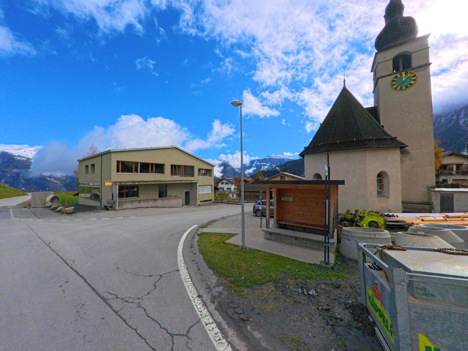 An einer leichten Strassenkurve stehen die Kirche mit Uhrturm und eine Bushaltestelle im Dorfzentrum von Urmein (7427, Kanton Graubünden) vor alpiner Kulisse.