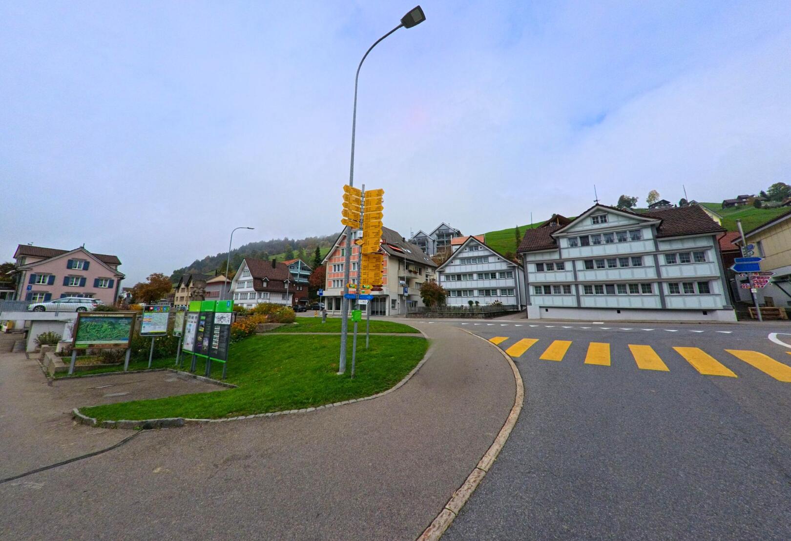 Vor traditionellen Hausern in Urnaesch (9107, Kanton Appenzell Ausserrhoden) fuhrt ein breiter Weg an einer Wegweisergruppe und einem Zebrastreifen vorbei.