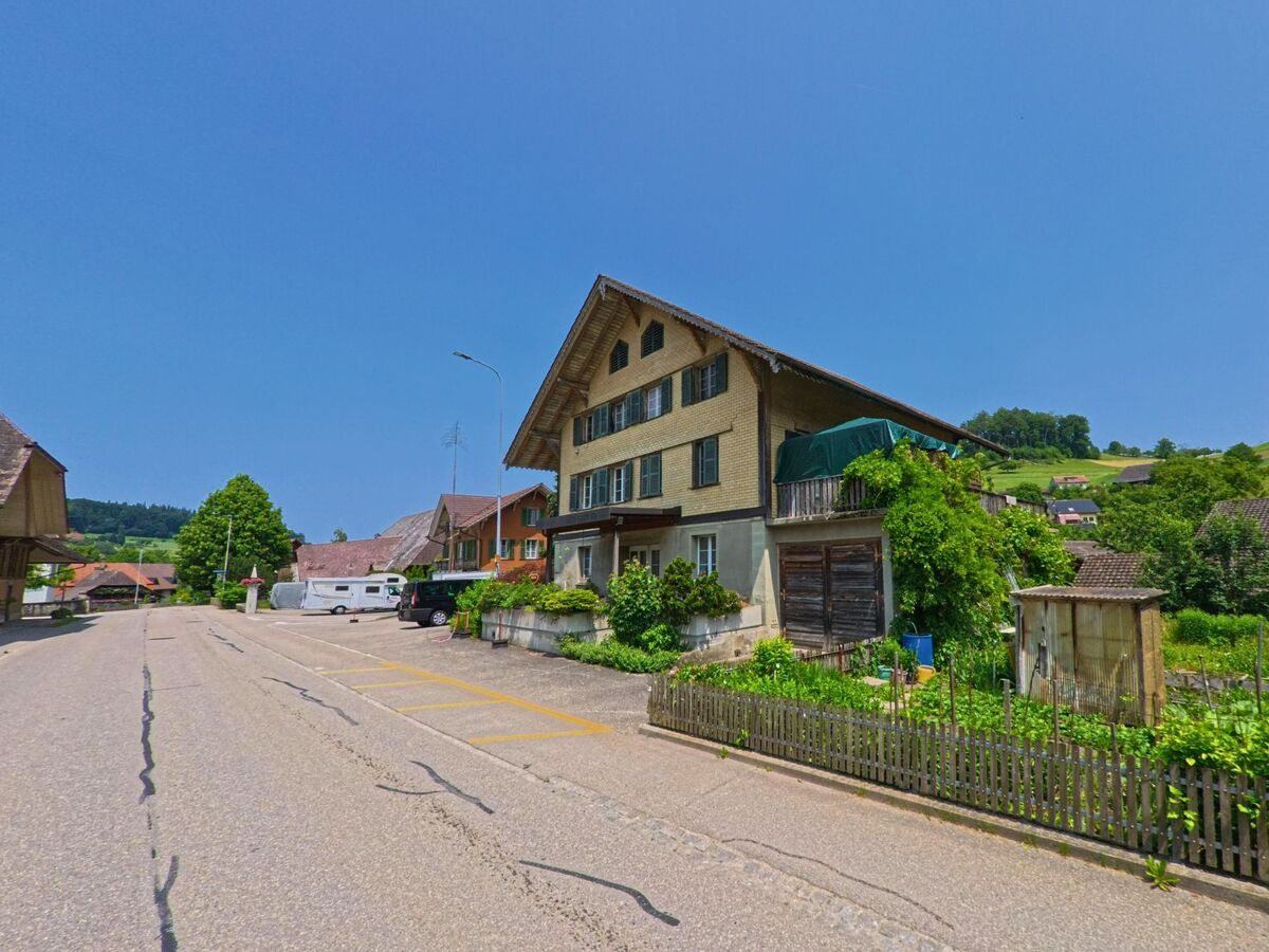 In Ursenbach (4937, Kanton Bern) reiht sich ein altes Wohnhaus mit Holzlaube und Gartenanlage entlang der ruhigen Dorfstrasse, umgeben von weiteren Hausern und Huegellandschaft.