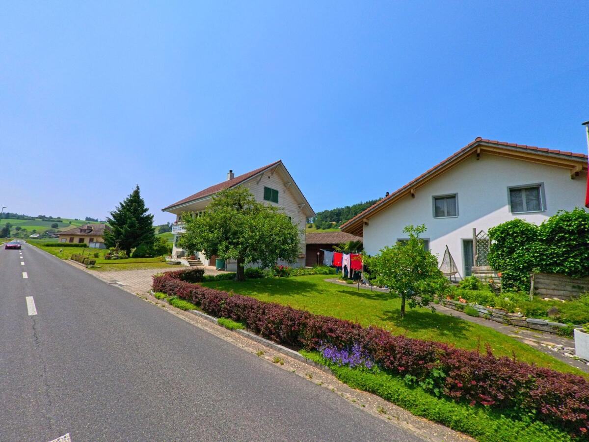 An einer Ausfallstrasse in Ursenbach (4937, Kanton Bern) reihen sich gepflegte Einfamilienhaeuser mit Vorgarten und Waescheleine vor einer sanften Huellandschaft.