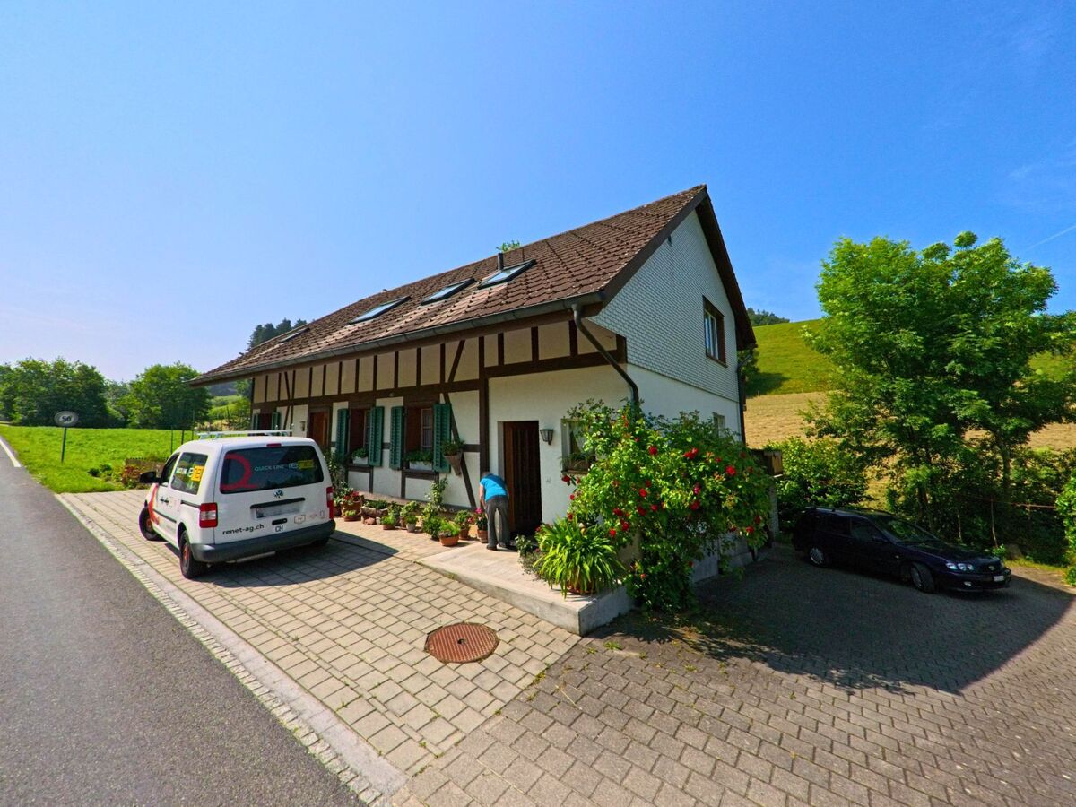 Ein Fachwerkhaus in Ursenbach (4937, Kanton Bern) liegt am Strassenrand mit blumengeschmücktem Eingangsbereich, parkenden Fahrzeugen und sanft ansteigendem Hügel im Hintergrund.