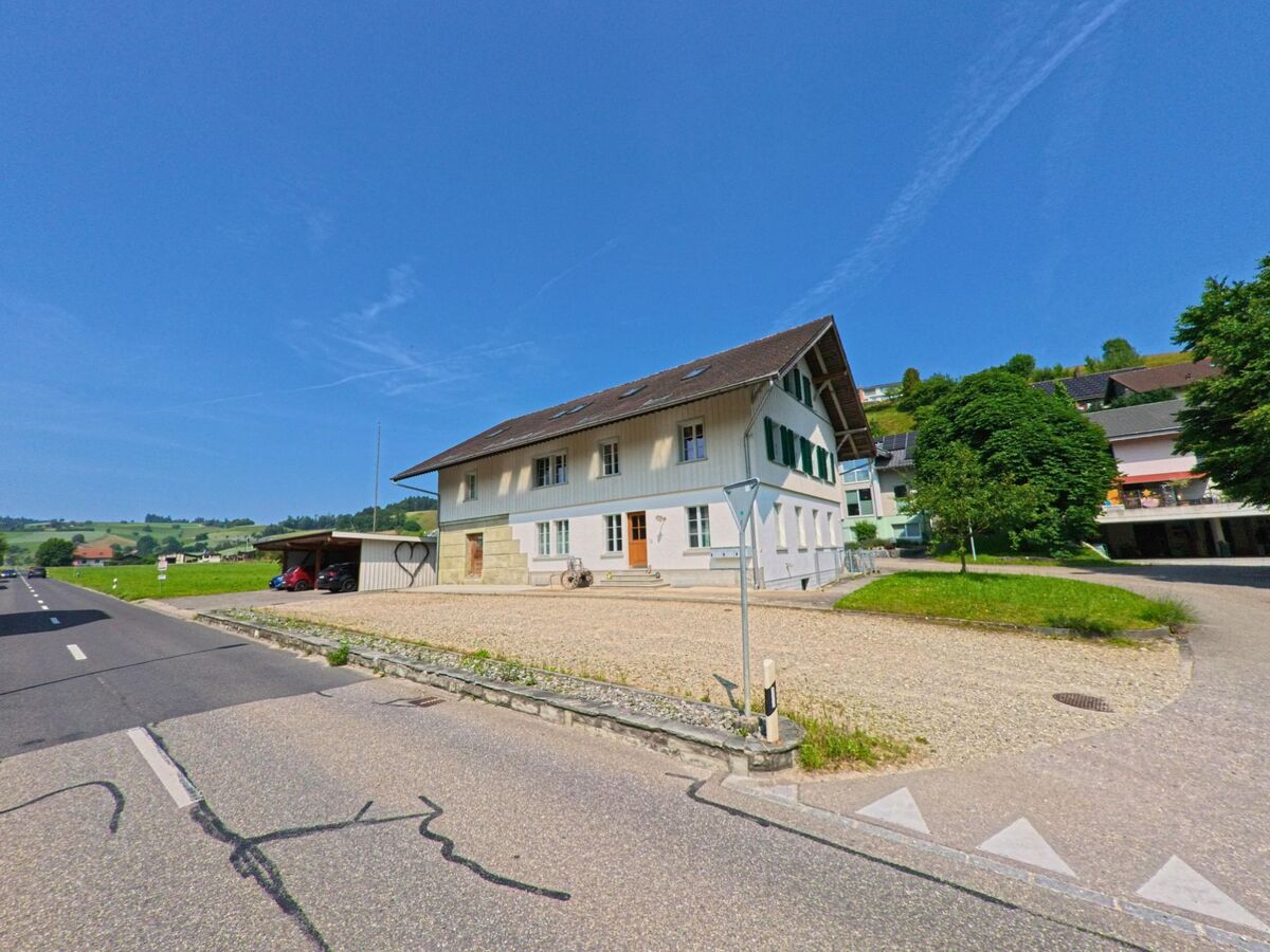 Am Ortsrand von Ursenbach (4937, Kanton Bern) steht ein helles Gebaeude mit gruenen Fensterlaeden und Kiesvorplatz direkt an der Hauptstrasse mit angrenzendem Carport.