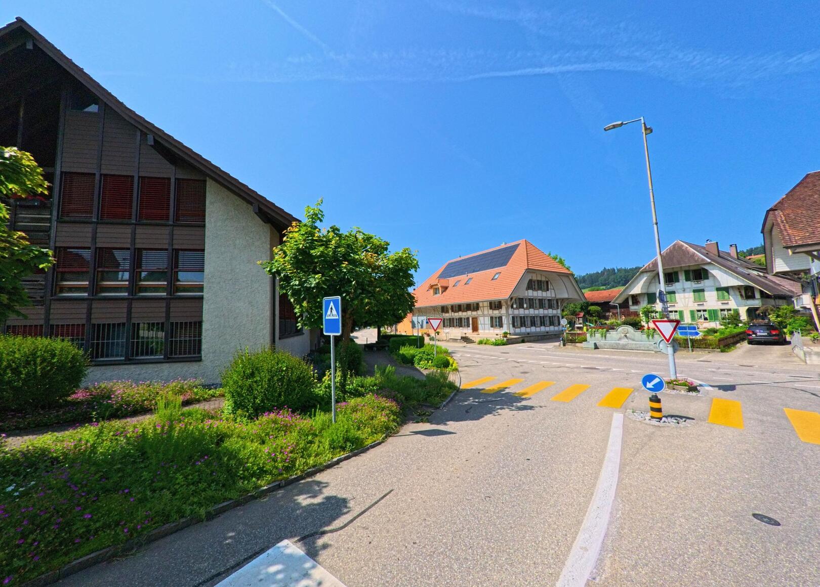 In Ursenbach (4937, Kanton Bern) kreuzen sich an einem sonnigen Tag mehrere Strassen zwischen traditionellen Wohnhäusern und einem modernen Schulgebäude mit gepflegter Umgebung.