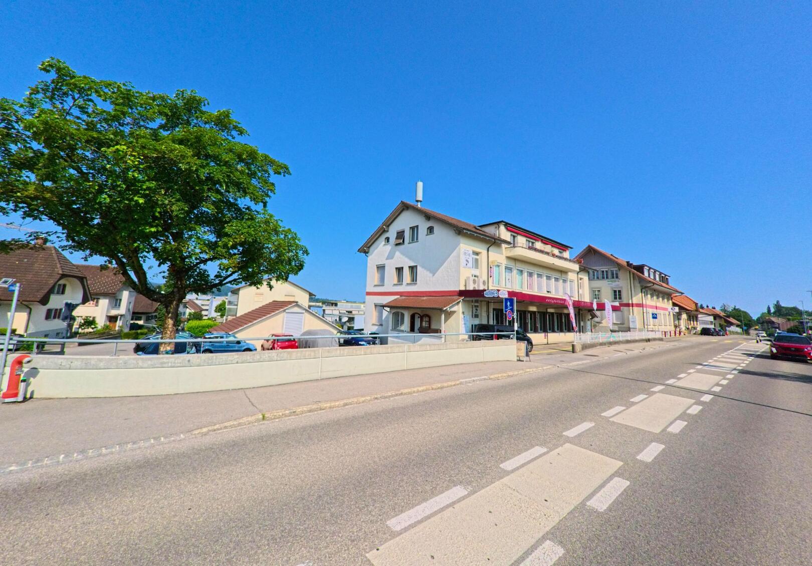 Entlang einer breiten Strasse mit Zebrastreifen stehen Wohn- und Geschäftsgebäude in Urtenen-Schönbühl (3322, Kanton Bern) unter klarem blauem Himmel nebeneinander.