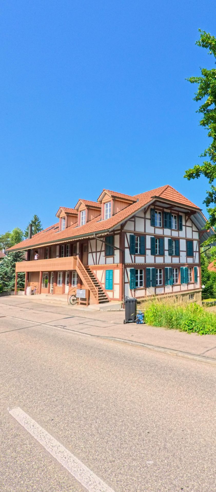Ein grosszügiges Fachwerkgebäude mit grünen Fensterläden und vorgelagerter Treppe steht an einer ruhigen Strasse in Urtenen-Schönbühl (3322, Kanton Bern) unter klarem Sommerhimmel.