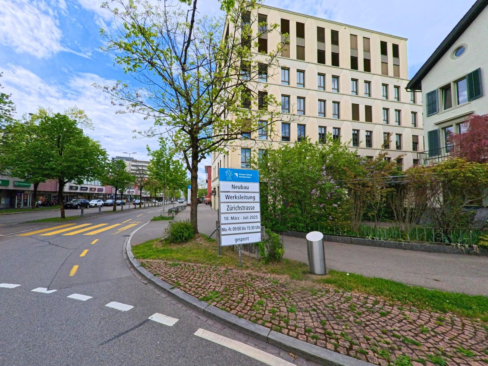 In Uster (8610, Kanton Zürich) verläuft die Zürichstrasse zwischen modernen Gebäuden und Bäumen, flankiert von einem Hinweis auf Bauarbeiten.