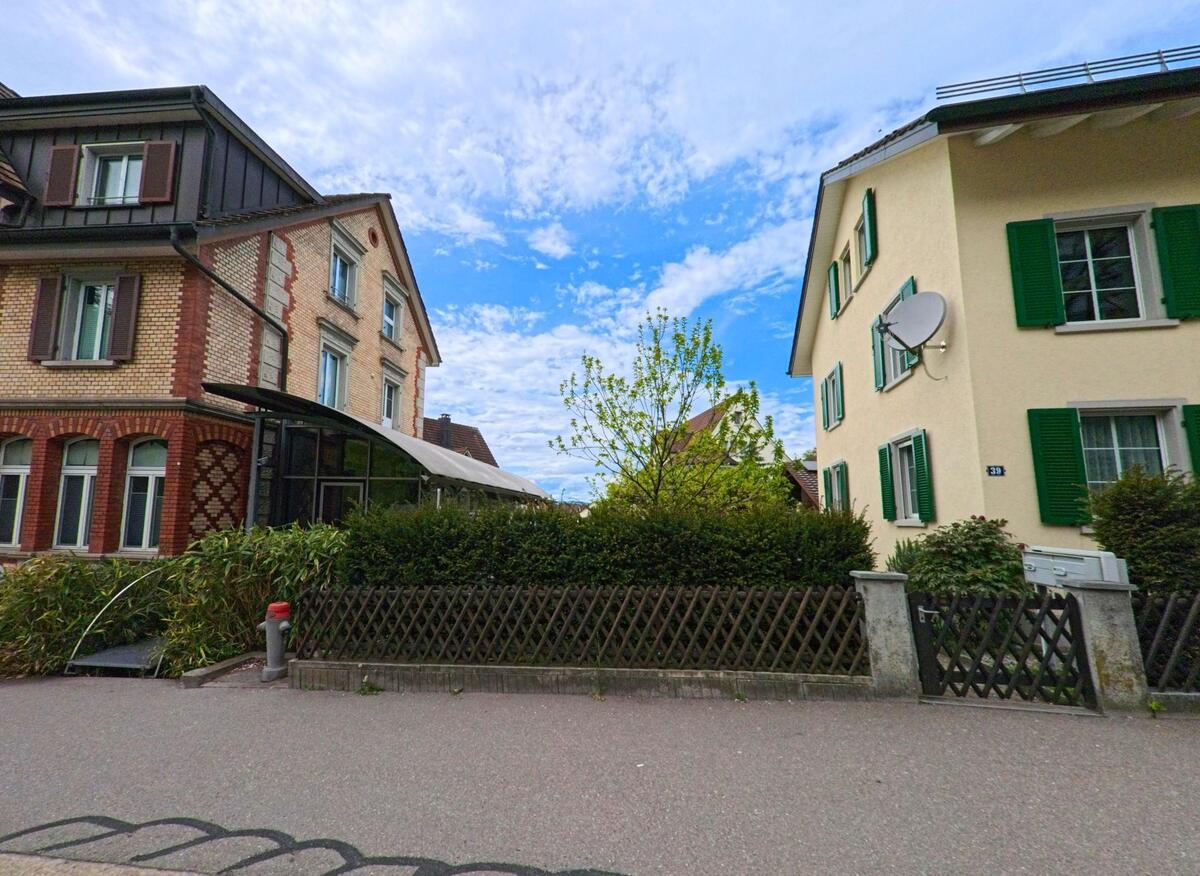 Zwei gepflegte Mehrfamilienhäuser in Uster (8610, Kanton Zürich) stehen sich gegenüber, getrennt durch einen schmalen Gartenstreifen mit Hecken und Zäunen.