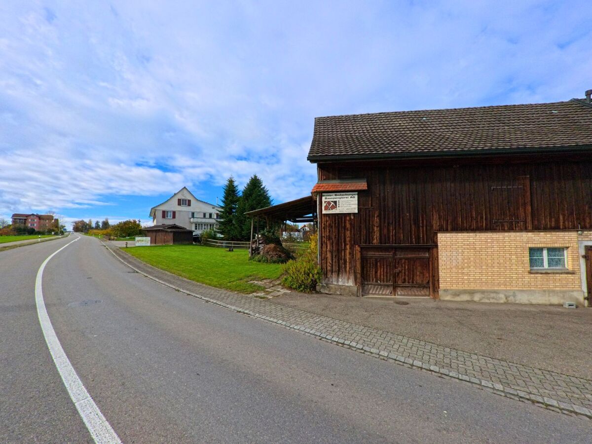 Am Rand einer Durchgangsstrasse in Uttwil (8592, Kanton Thurgau) steht eine Holzscheune mit angrenzender Gruenflaeche und Wohnhaus im Hintergrund unter weit gespanntem Himmel.