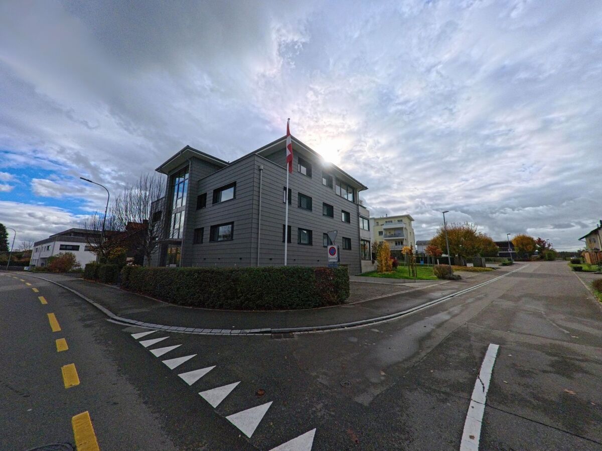 An einer Ecke in Uttwil (8592, Kanton Thurgau) steht ein modernes Verwaltungsgebaeude mit Schweizer Flagge an einer ruhigen Quartierstrasse unter dicht bewolktem Himmel.
