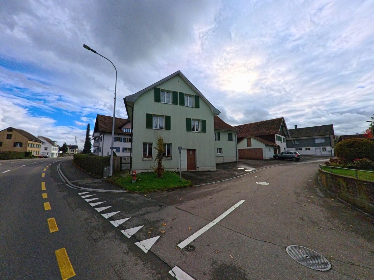 An einer Strassenkreuzung in Uttwil (8592, Kanton Thurgau) steht ein helles Wohnhaus mit Gruenflaeche, umgeben von ruhigen Quartierstrassen unter wechselhaftem Himmel.
