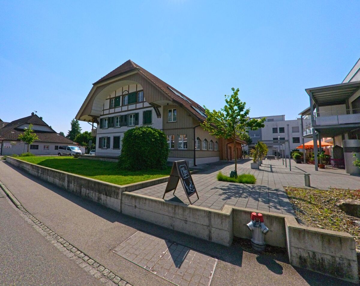 In Utzenstorf (3427, Kanton Bern) steht ein traditionelles Wohnhaus mit ausladendem Dachgiebel direkt neben einem modernen Platz mit kleinen Bäumen und Cafébetrieb.