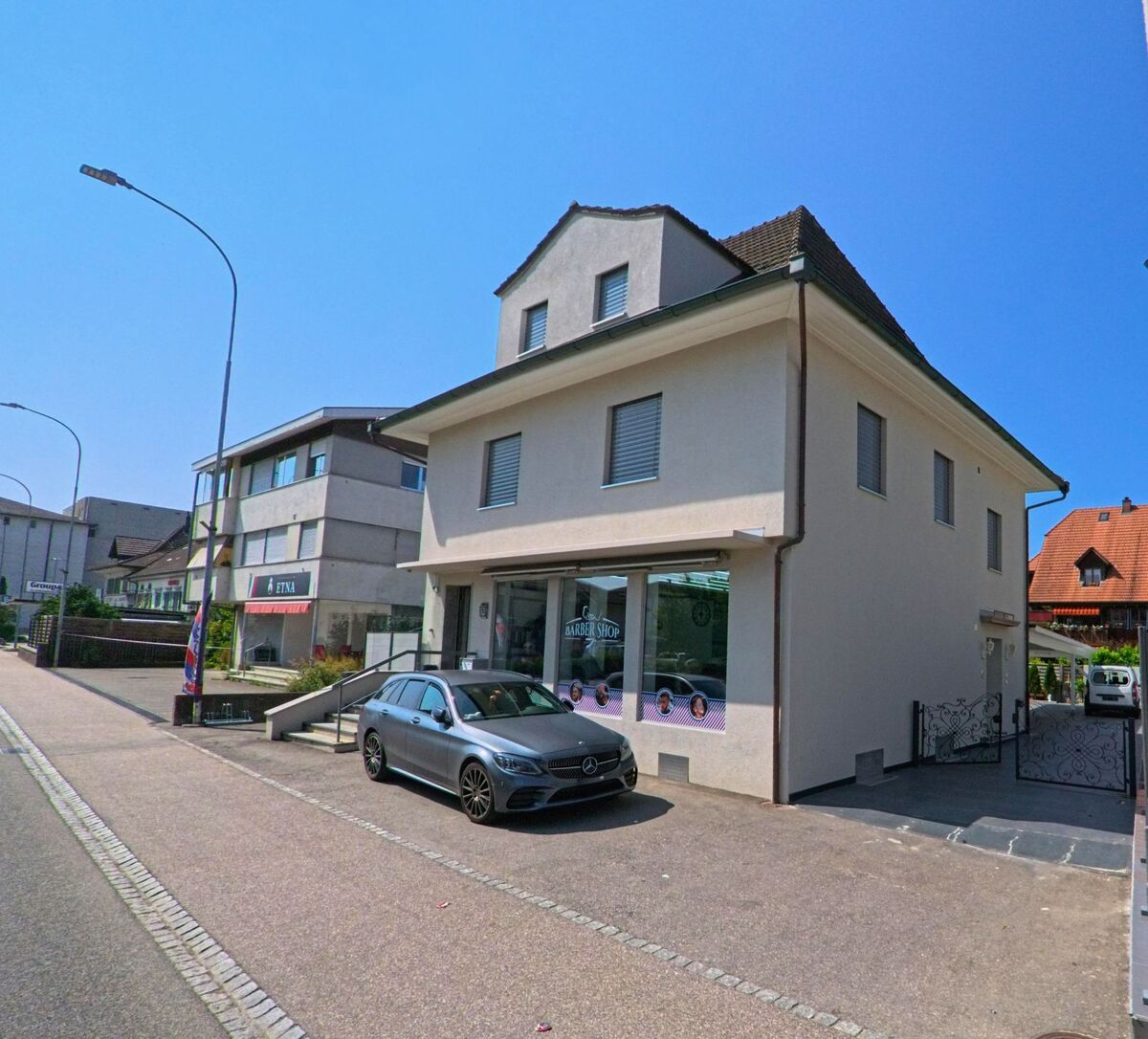 In Utzenstorf (3427, Kanton Bern) befindet sich ein modernes Geschäftshaus mit Barber Shop an einer Hauptstrasse, flankiert von weiteren Gewerbebauten.