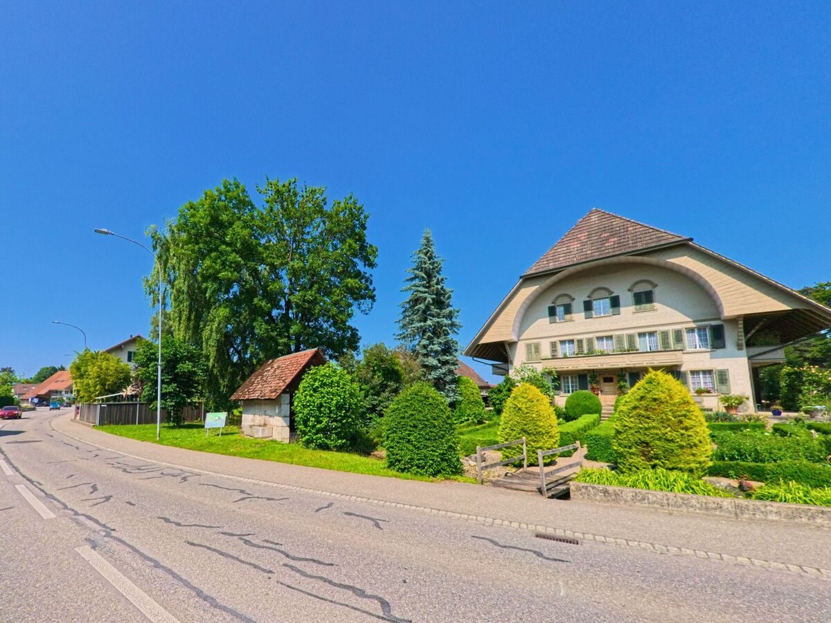 Entlang der Hauptstrasse in Utzenstorf (3427, Kanton Bern) steht ein grosses Bauernhaus mit weit ausladendem Dach, umgeben von einem gepflegten Garten mit Ziergehölzen.