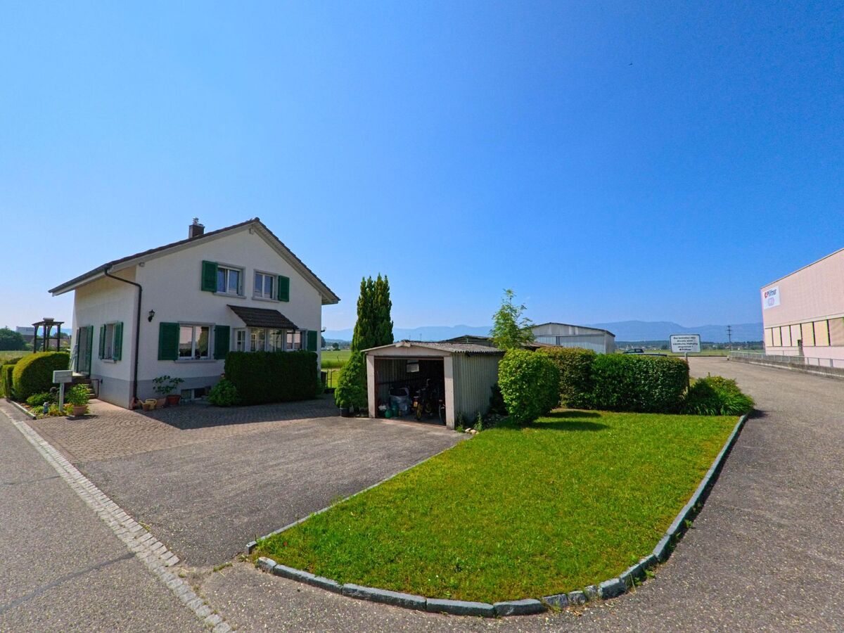 Am Rand eines Gewerbegebiets in Utzenstorf (3427, Kanton Bern) steht ein Einfamilienhaus mit gepflegtem Vorplatz und kleiner Garage, dahinter öffnet sich der Blick auf Felder und Berge.