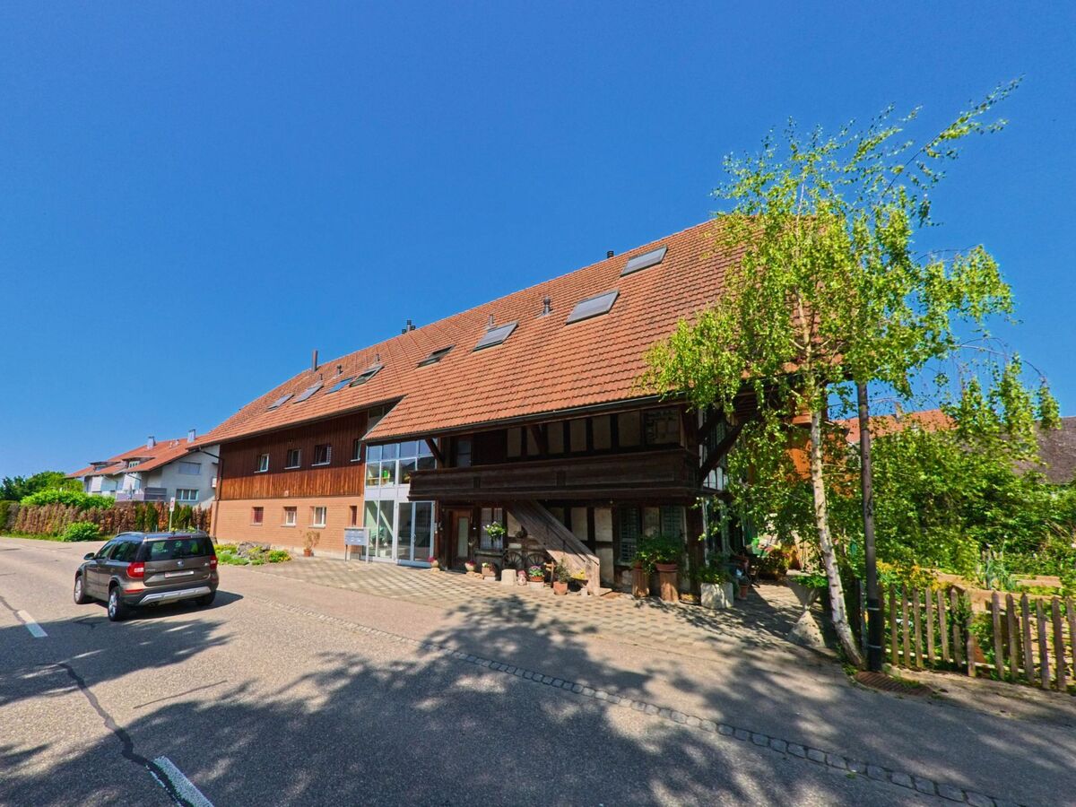 In Utzenstorf (3427, Kanton Bern) fährt ein Auto an einem langgezogenen Wohngebäude mit Holzfassade und traditionellem Eingang vorbei, das sich harmonisch in die Siedlungsstruktur einfügt.