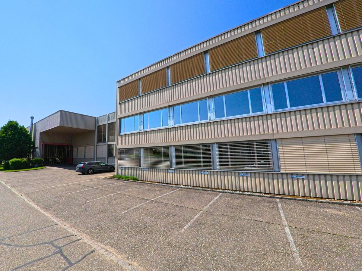 Ein modernes Verwaltungsgebäude mit vertikaler Fassadenstruktur steht in Utzenstorf (3427, Kanton Bern) hinter einem fast leeren Parkplatz bei klarem Sommerwetter.