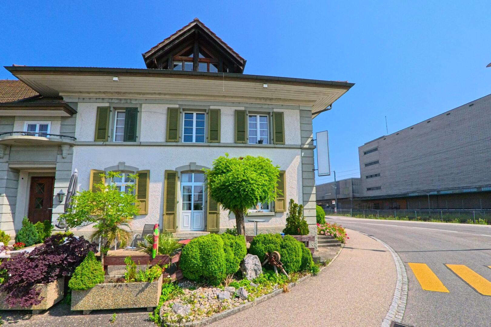 In Utzenstorf (3427, Kanton Bern) steht ein gepflegtes Wohnhaus mit gruener Fensterlaeden direkt an einer Strasse, die entlang eines grossen Industriegebäudes verläuft.