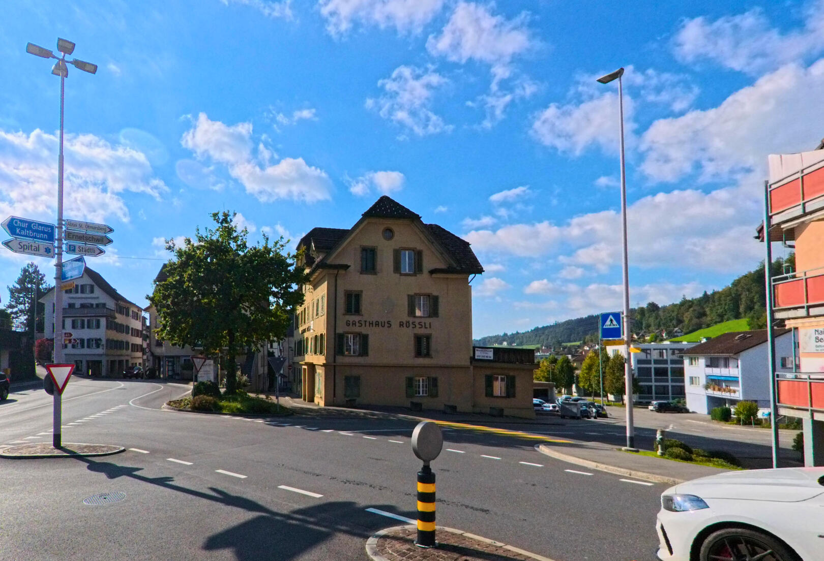 An einer verkehrsreichen Kreuzung in Uznach (8730, Kanton St. Gallen) steht ein historisches Gebäude neben Fahrbahnen, Wegweisern und moderner Strassenbeleuchtung unter blauem Himmel.