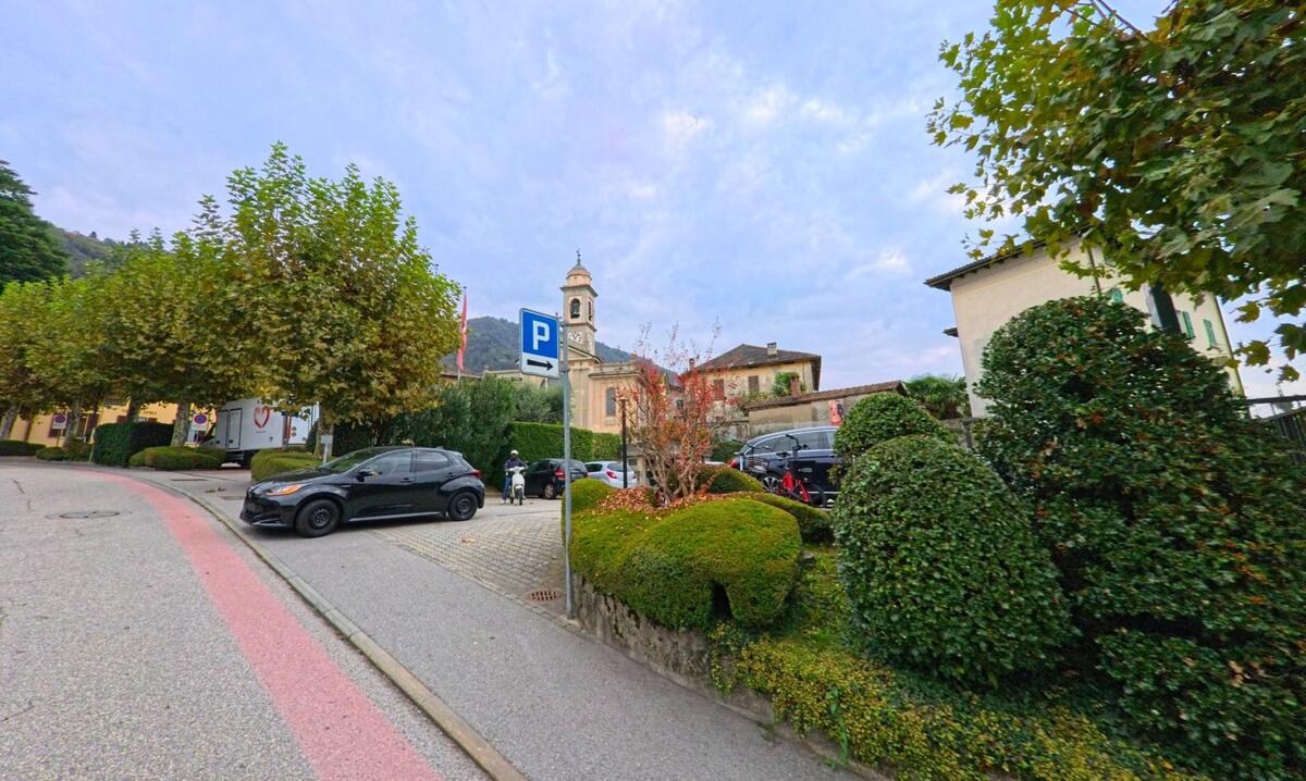 Ein gepflegter Platz in Vacallo (6833, Kanton Tessin) mit Bäumen, geparkten Autos und Blick auf die Kirche mit ihrem Glockenturm.