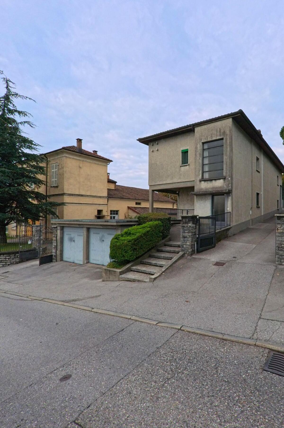 Zwei Wohnhäuser in Vacallo (6833, Kanton Tessin) liegen nebeneinander, verbunden durch eine gepflasterte Einfahrt mit Garagentoren und gepflegter Hecke.