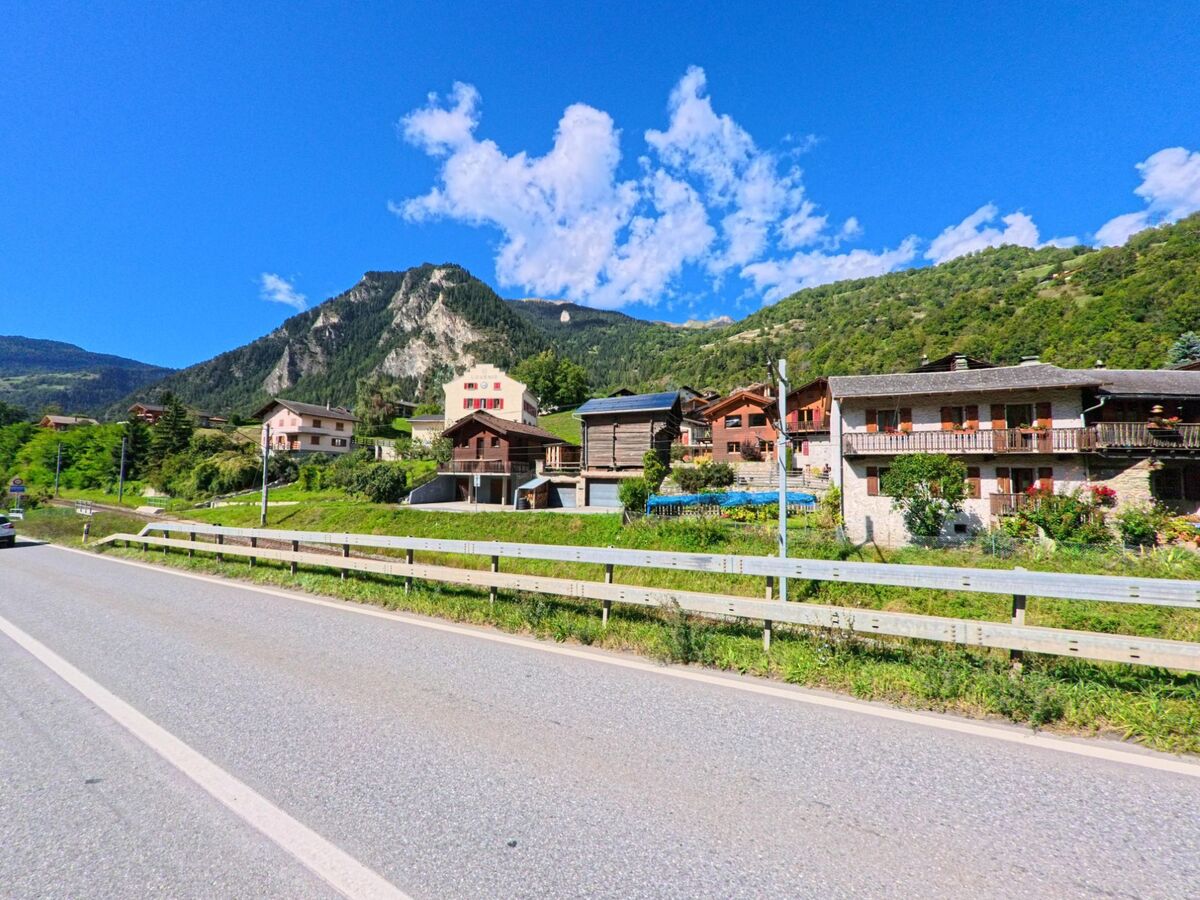 Eine Dorfzeile mit Chalets und Wohnhaeusern liegt oberhalb einer Strasse in Val de Bagnes (1934, Kanton Wallis) vor steiler Bergkulisse.
