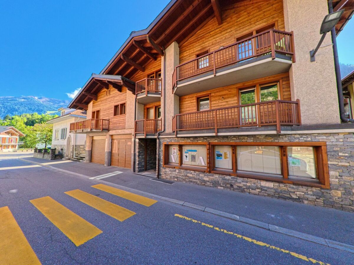 Ein mehrgeschossiges Wohn- und Geschaeftsgebaeude mit Holzfassade und Balkonen steht an einer Dorfstrasse in Val de Bagnes (1934, Kanton Wallis).