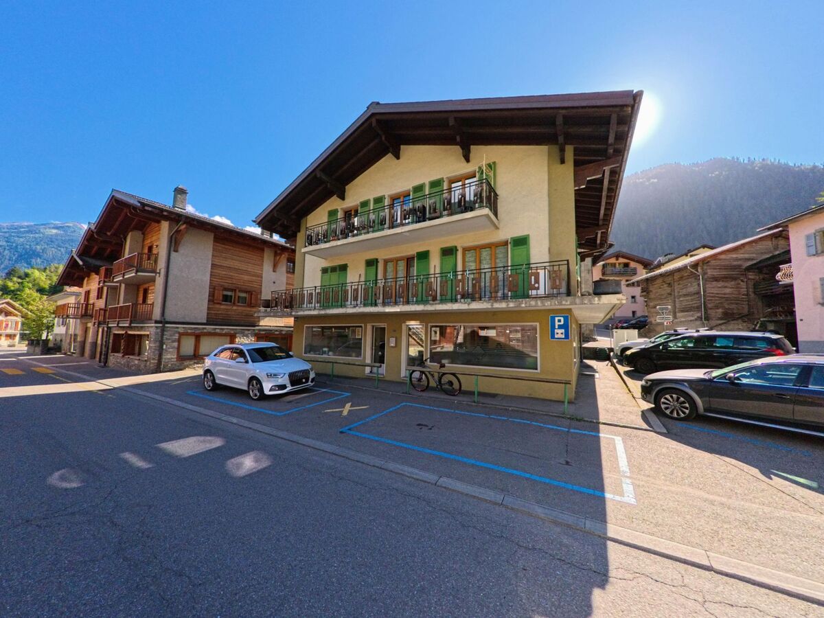 Ein Wohn- und Geschaeftshaus mit Balkonen und Parkplaetzen steht an einer Dorfstrasse in Val de Bagnes (1934, Kanton Wallis) bei klarem Wetter.
