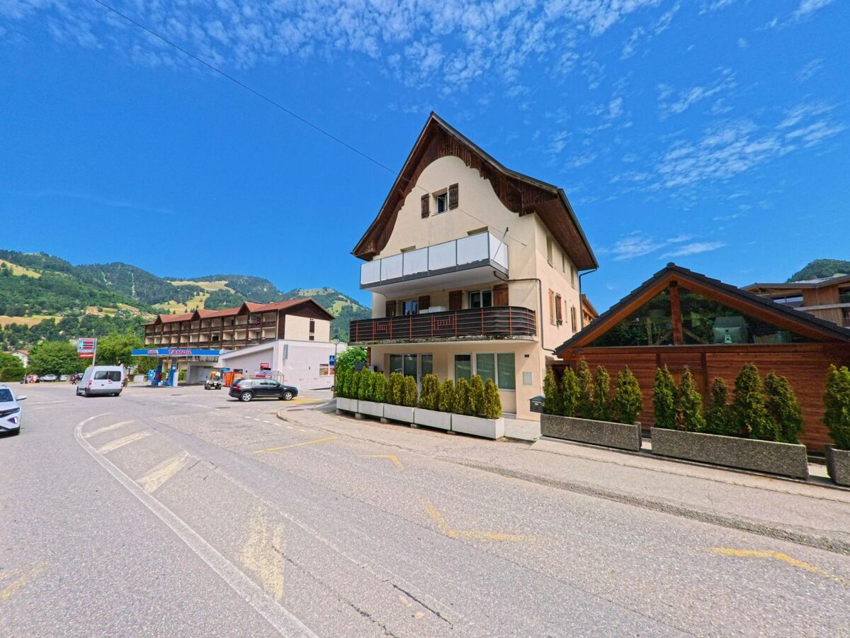 Entlang einer Dorfstrasse in Val-de-Charmey (1637, Kanton Freiburg) stehen ein Wohnhaus mit Balkonen sowie weitere Gebäude vor den grünen Hängen der Umgebung.