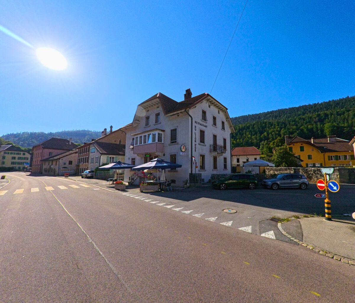 An einer Kreuzung in Val-de-Travers (2115, Kanton Neuenburg) steht ein mehrgeschossiges Gebaeude mit Restaurant und Aussenterrasse, umgeben von weiteren Haeusern und bewaldeten Huegeln.