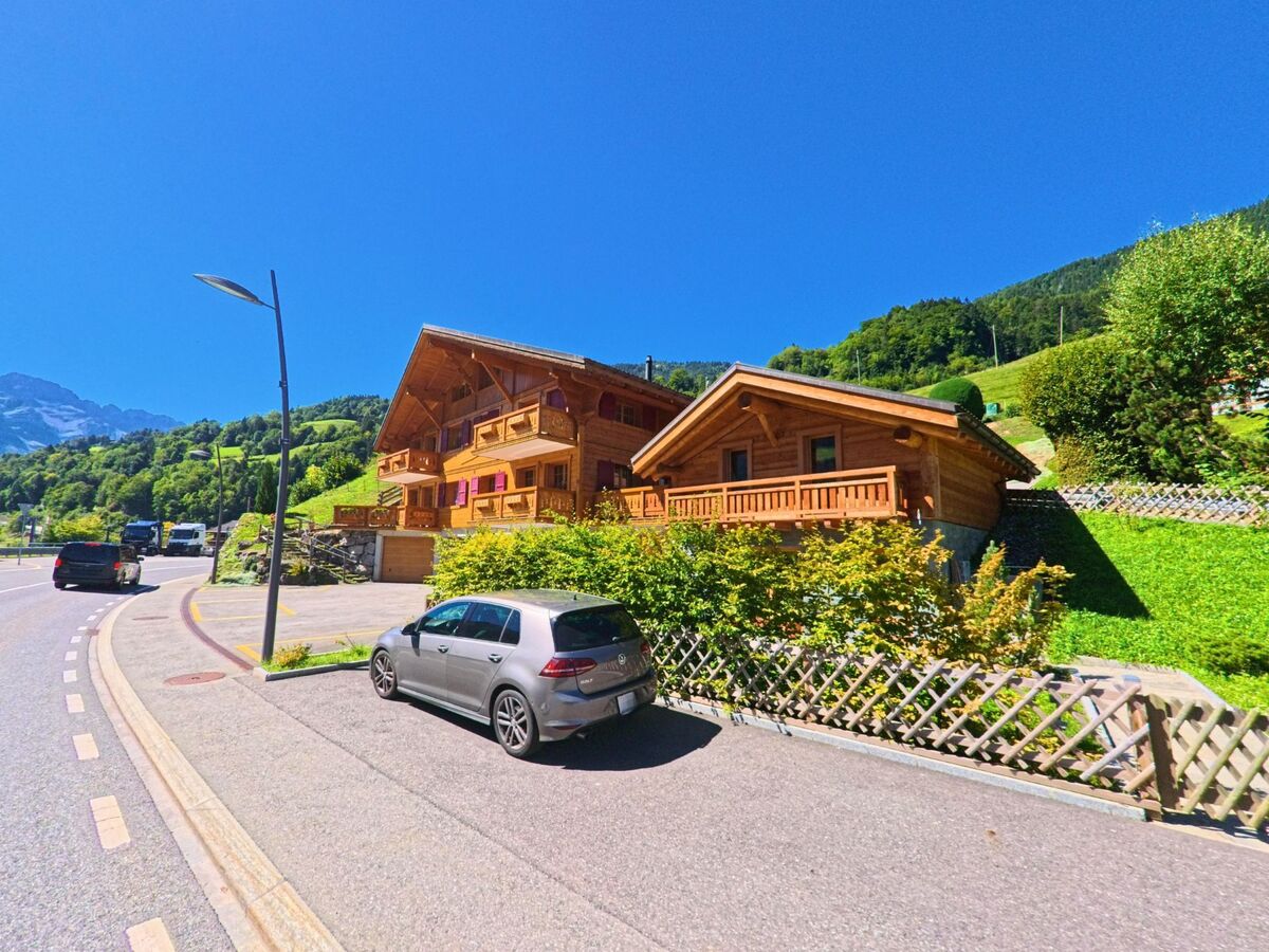 Zwei moderne Holzhäuser in Chalet-Bauweise stehen am Strassenrand von Val-d'Illiez (1873, Kanton Wallis) vor der Kulisse grüner Berghänge.