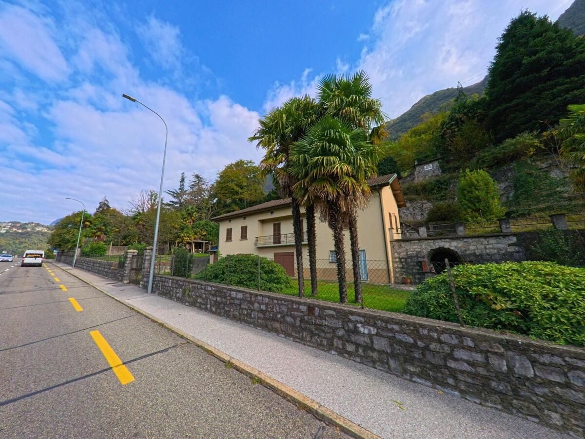 Ein Haus mit hellbeiger Fassade und mehreren Palmen steht in Val Mara (6818, Kanton Tessin) direkt an einer breiten Strasse mit Bergkulisse im Hintergrund.