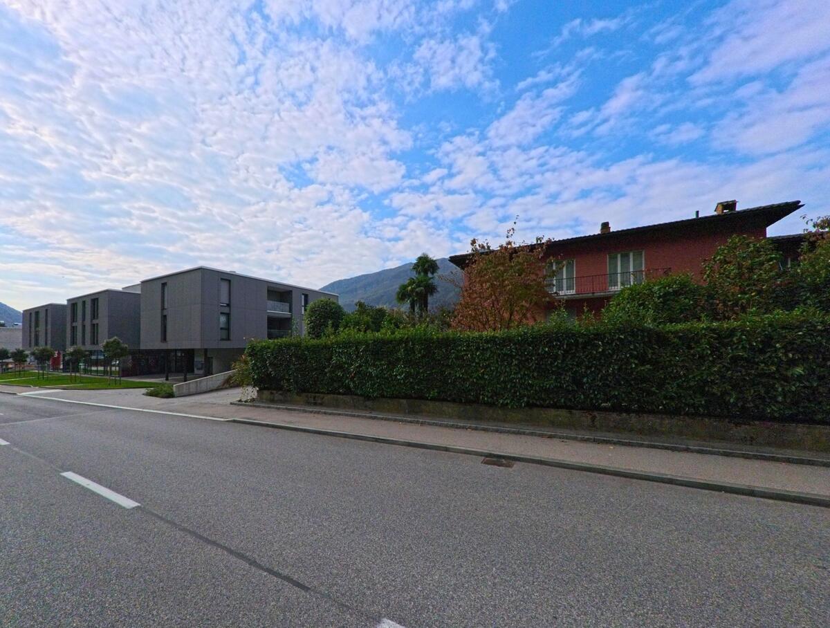 Moderne Wohnbauten und ein rotes Mehrfamilienhaus praegen in Val Mara (6818, Kanton Tessin) eine gepflegte Strasse mit Blick auf die umliegenden Berge.