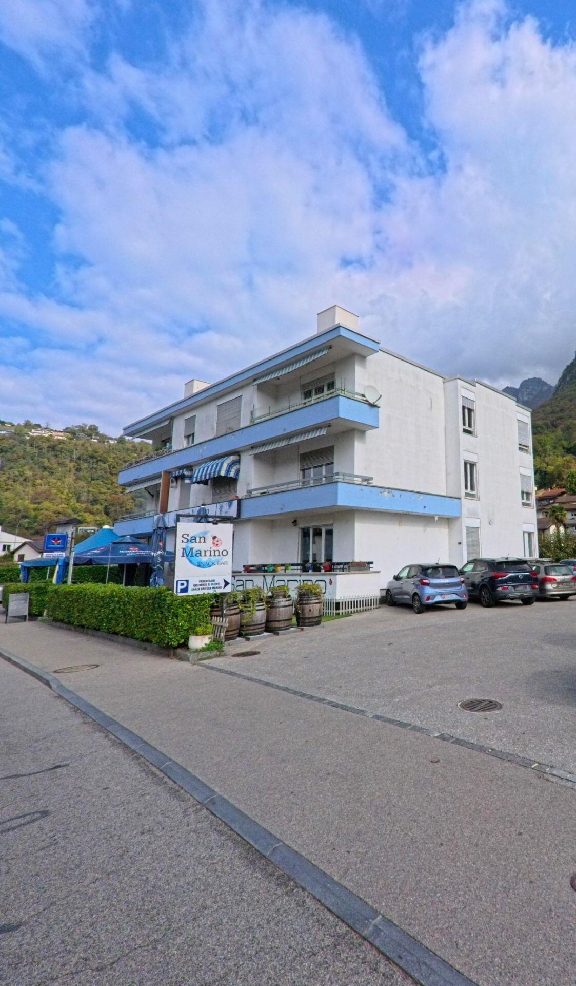 Ein helles Gebaeude mit Balkonen und dem Restaurant San Marino steht in Val Mara (6818, Kanton Tessin) an einer Strasse mit Parkplatz und Bergkulisse.