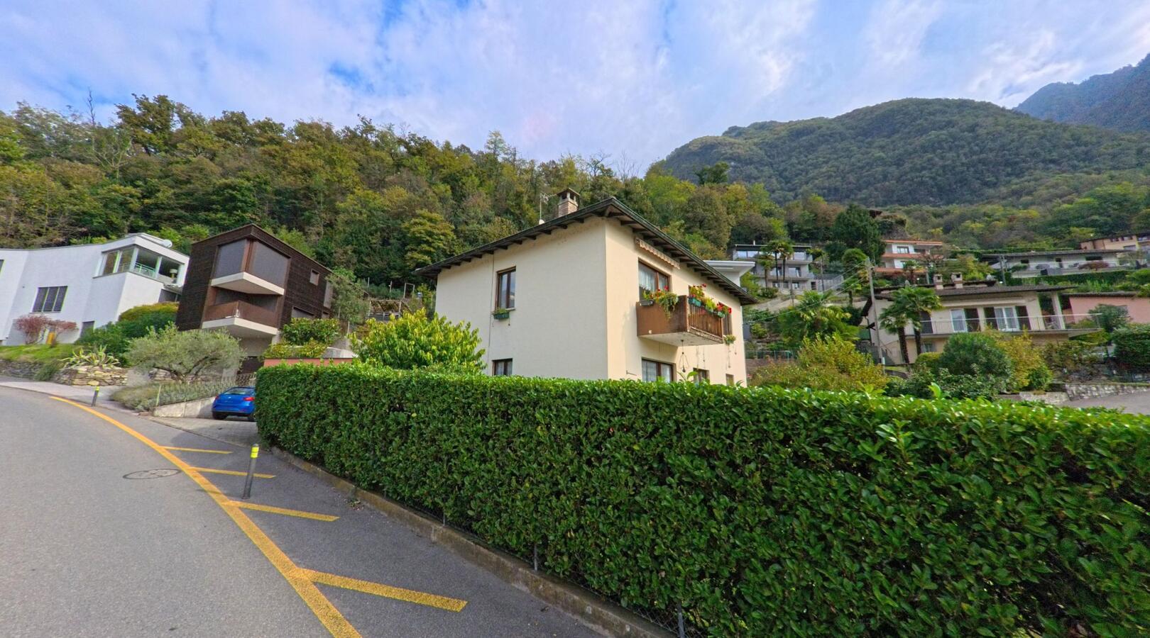 In Val Mara (6818, Kanton Tessin) stehen entlang einer kurvigen Strasse gepflegte Wohnhaeuser mit Balkonen und dichter Hecke vor bewaldetem Hang.
